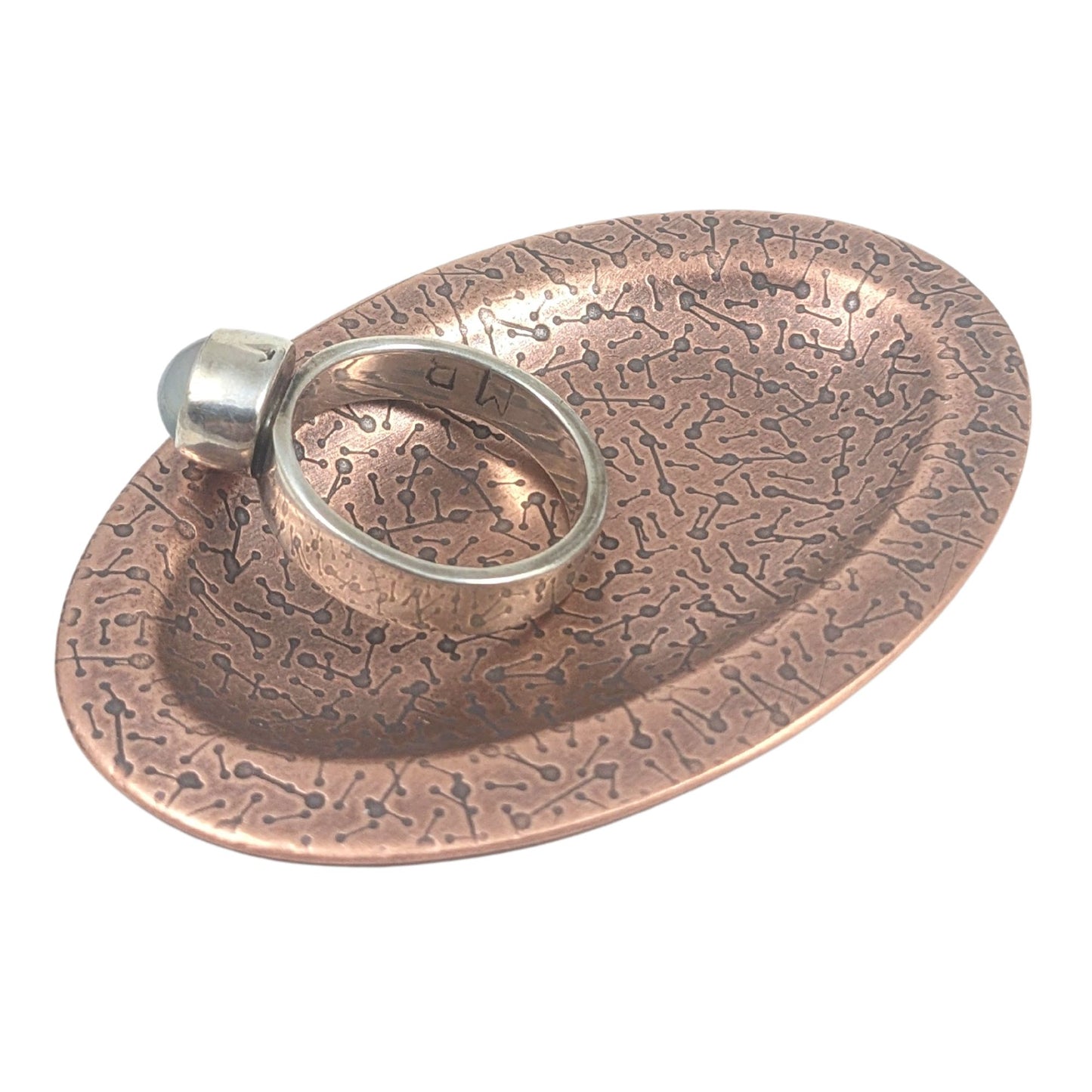 Oval ring dish made of copper. The dish has a shallow bowl with a lip around the edge. It is covered in a pattern meant to represent a meteor shower. The pattern is impressed into the copper and is short lines with small solid circles at the end. The design is oxidized, which means the impressed parts are dark, almost black. Staged with a ring.