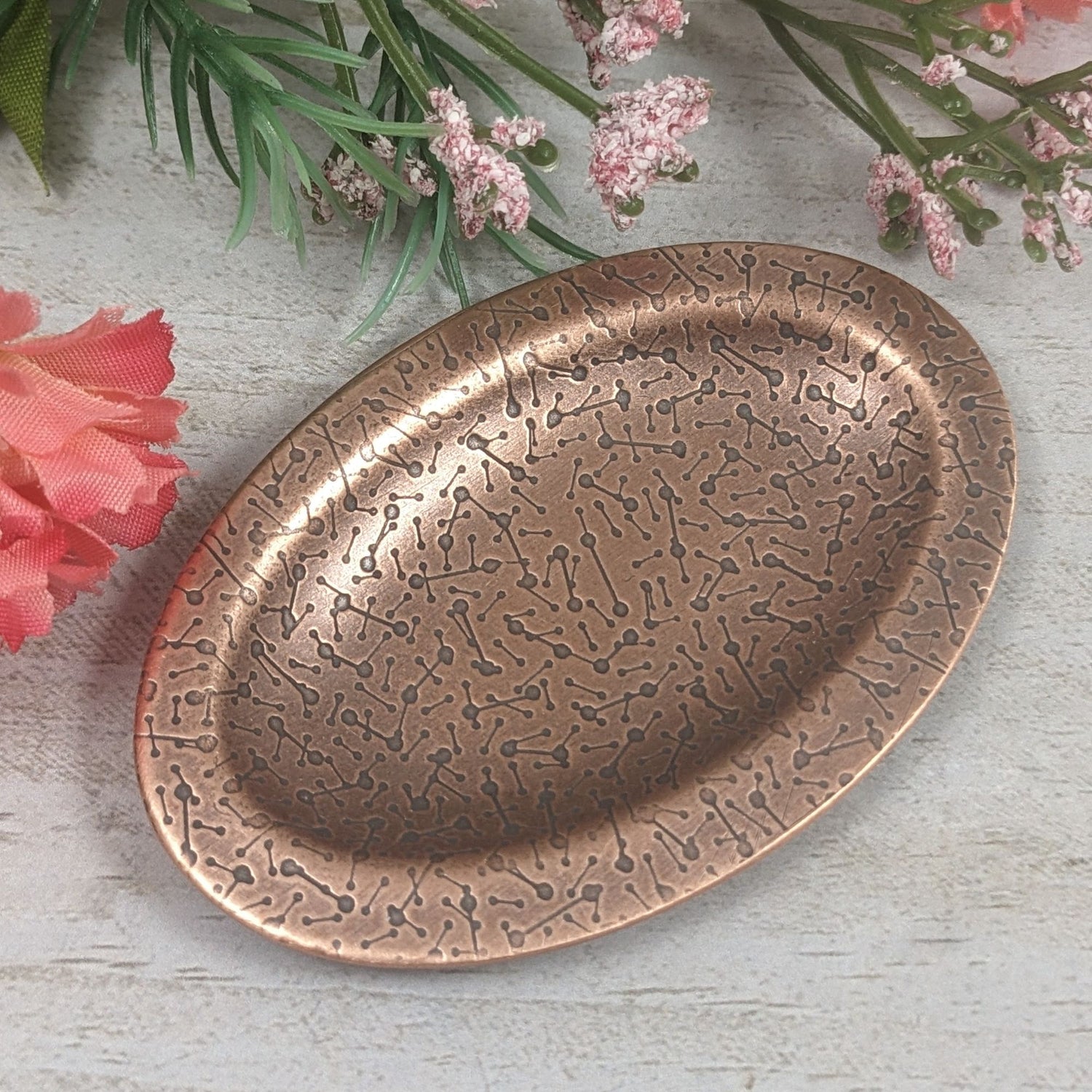Oval ring dish made of copper. The dish has a shallow bowl with a lip around the edge. It is covered in a pattern meant to represent a meteor shower. The pattern is impressed into the copper and is short lines with small solid circles at the end. The design is oxidized, which means the impressed parts are dark, almost black. Stages with flowers in background.
