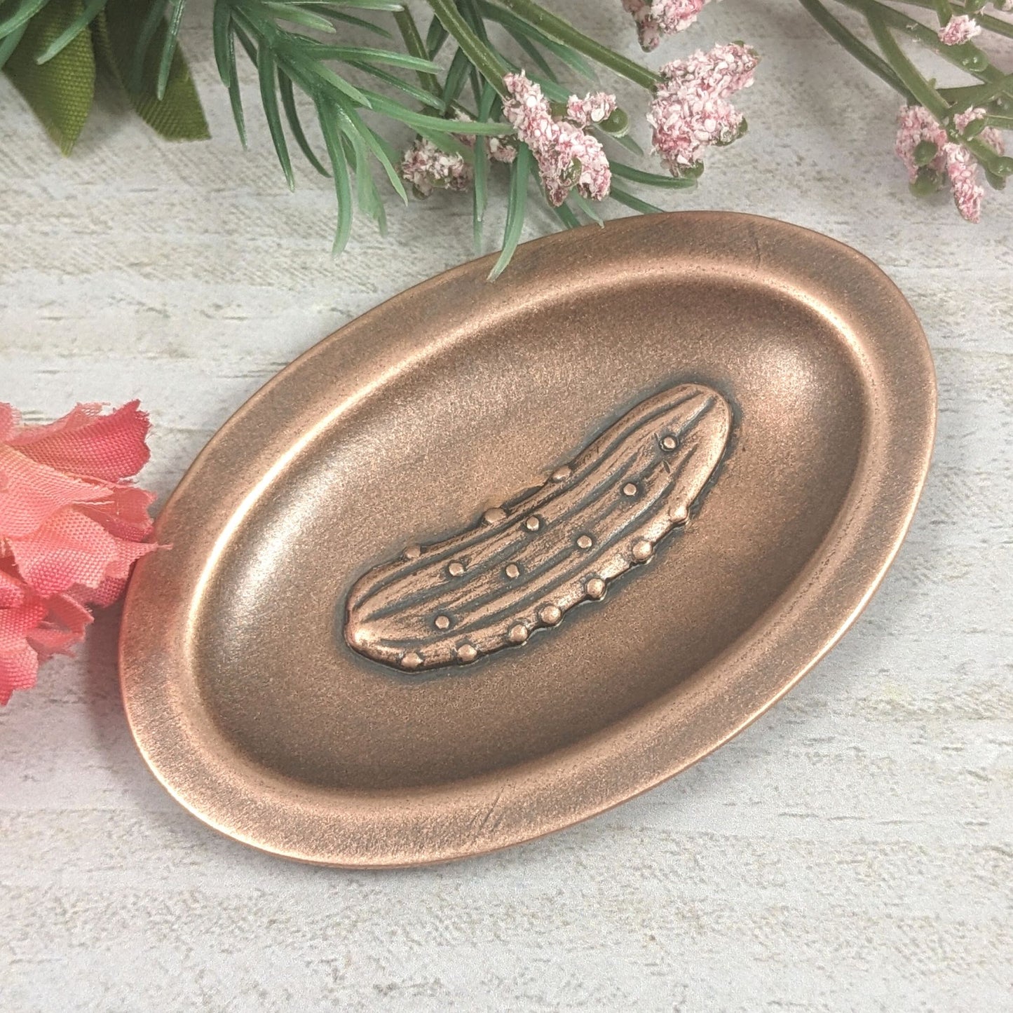 Oval ring dish made of copper. The dish has a shallow bowl with a lip around the edge. Centered on the bottom of the dish is a three-dimensional design of a pickle, also made of copper. The pickle is large and has stipples and ridges, just like a fresh from the garden pickle would have.  The design is oxidized, which means the recessed parts of the pickle are dark, almost black. Staged with flowers.