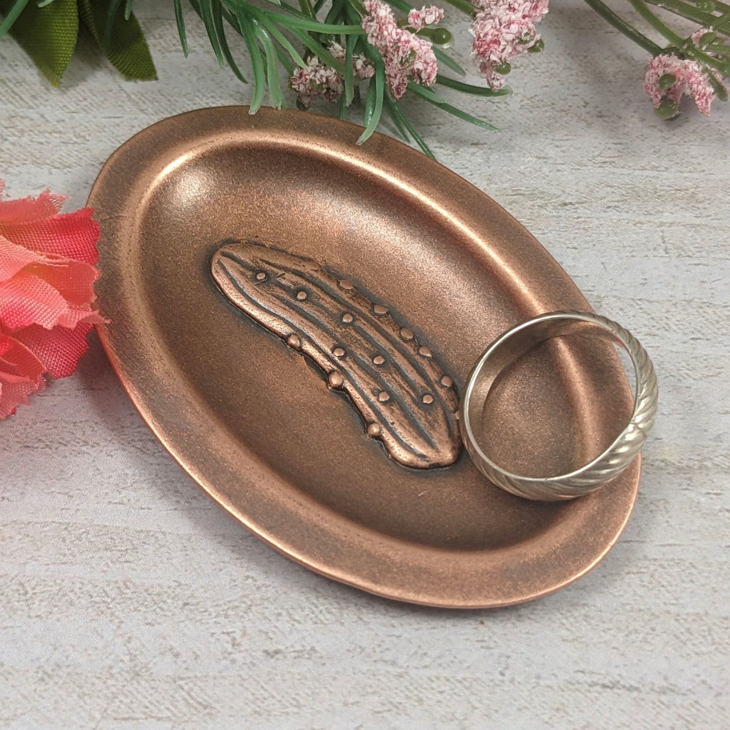 Oval ring dish made of copper. The dish has a shallow bowl with a lip around the edge. Centered on the bottom of the dish is a three-dimensional design of a pickle, also made of copper. The pickle is large and has stipples and ridges, just like a fresh from the garden pickle would have.  The design is oxidized, which means the recessed parts of the pickle are dark, almost black. Staged with a ring.