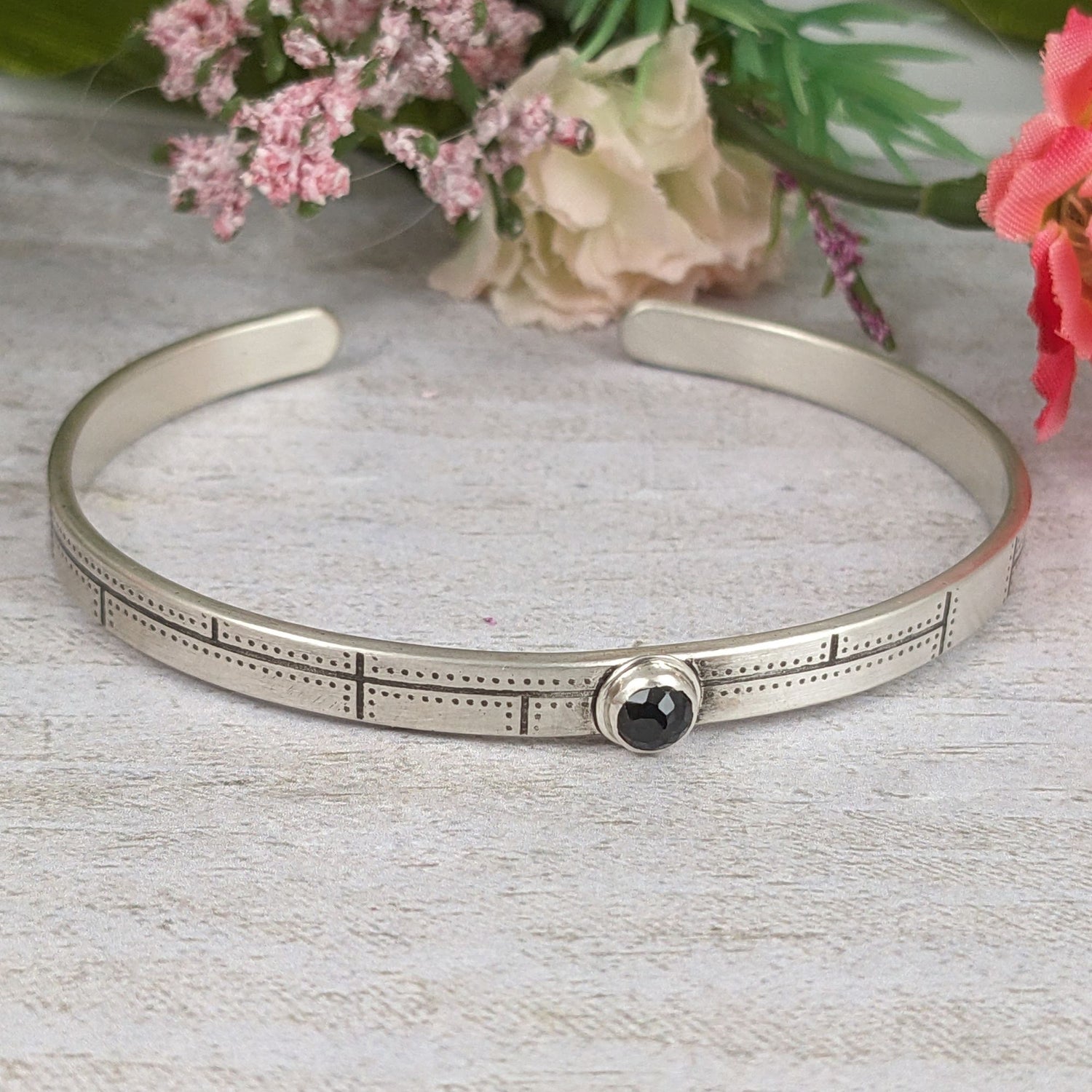 Narrow sterling silver cuff bracelet. The bracelet has a riveted panel design and features a rose cut black spinel gemstone. Staged with flowers in the background.