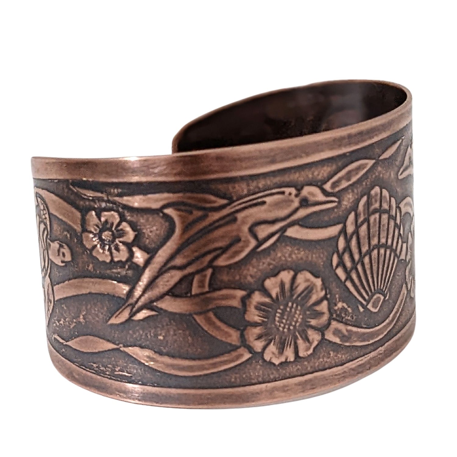 Wide tapered copper cuff bracelet with sea life design. Center is a seashell, with flowers and dolphins on either side.  Toward the ends of the cuff there are turtles, and a seaweed pattern is woven throughout the design.