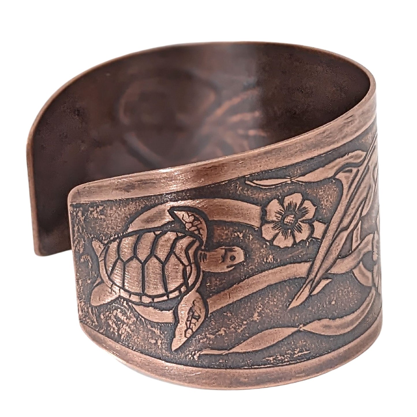 Wide tapered copper cuff bracelet with sea life design. Center is a seashell, with flowers and dolphins on either side.  Toward the ends of the cuff there are turtles, and a seaweed pattern is woven throughout the design.