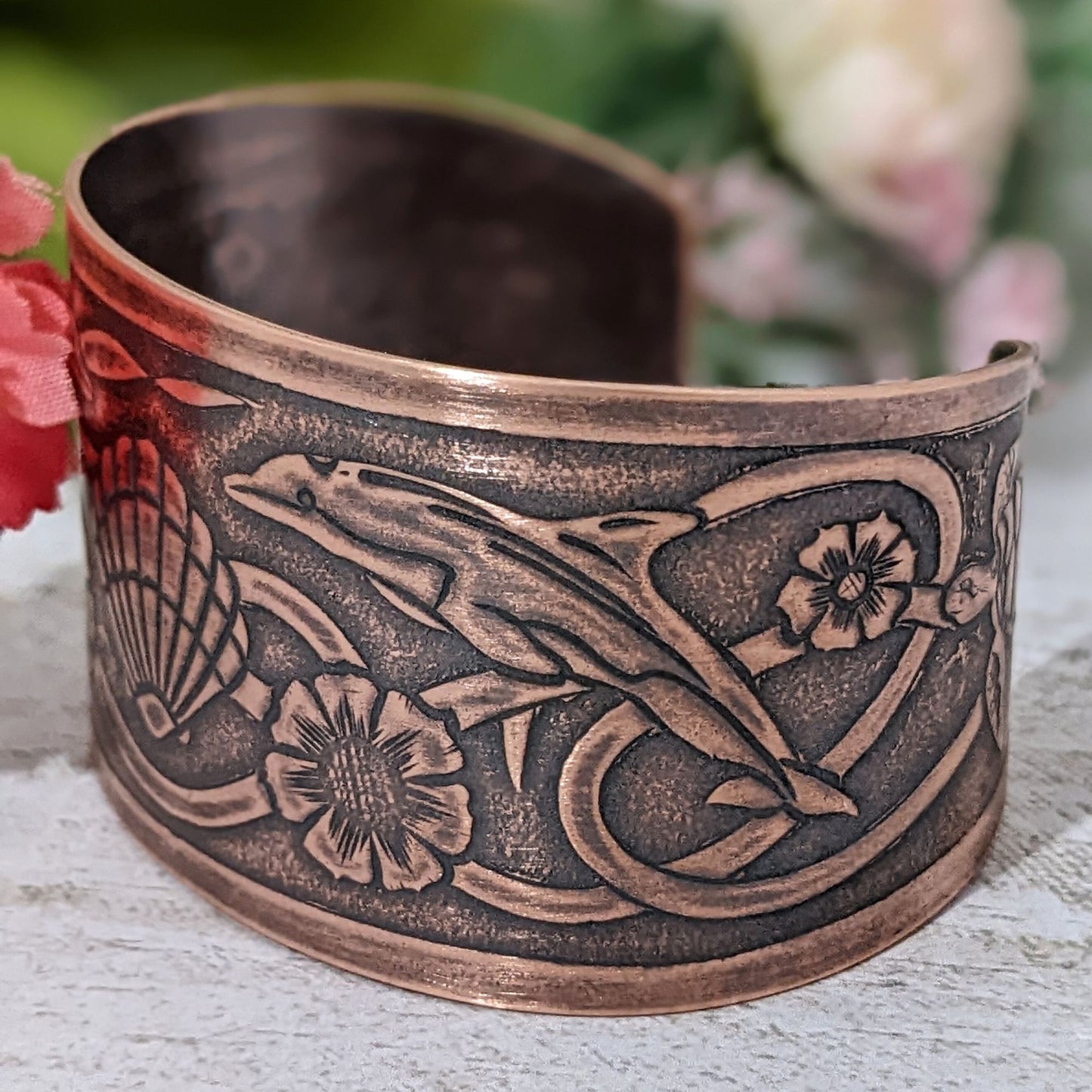 Wide tapered copper cuff bracelet with sea life design. Center is a seashell, with flowers and dolphins on either side.  Toward the ends of the cuff there are turtles, and a seaweed pattern is woven throughout the design.