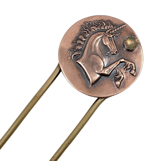 Brass hair fork with a copper design at the top. The copper is round with a raised impression of a unicorn. You can see the beard, detailed hair in the mane and on the ankles. The horn is spiraled. Above the nose there is a round gemstone meant to represent the moon.