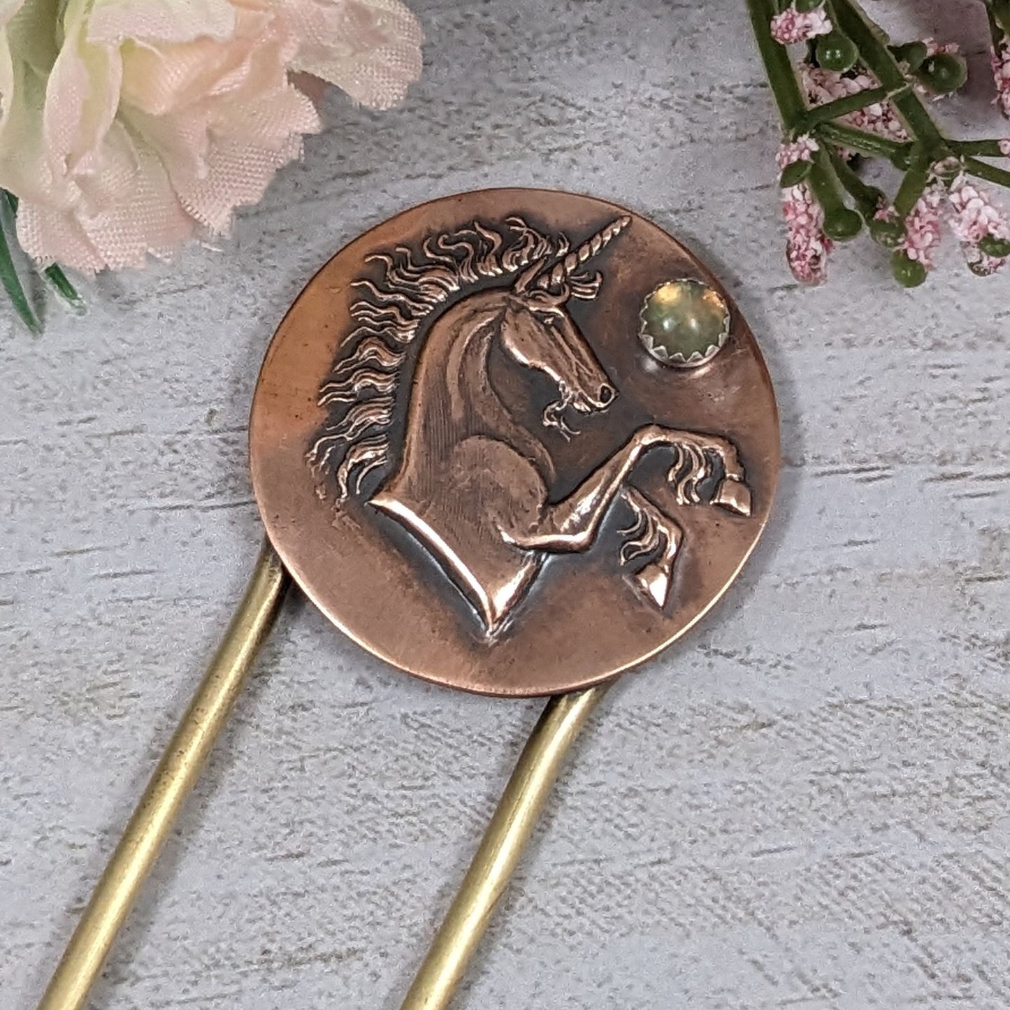 Brass hair fork with a copper design at the top. The copper is round with a raised impression of a unicorn. You can see the beard, detailed hair in the mane and on the ankles. The horn is spiraled. Above the nose there is a round gemstone meant to represent the moon.