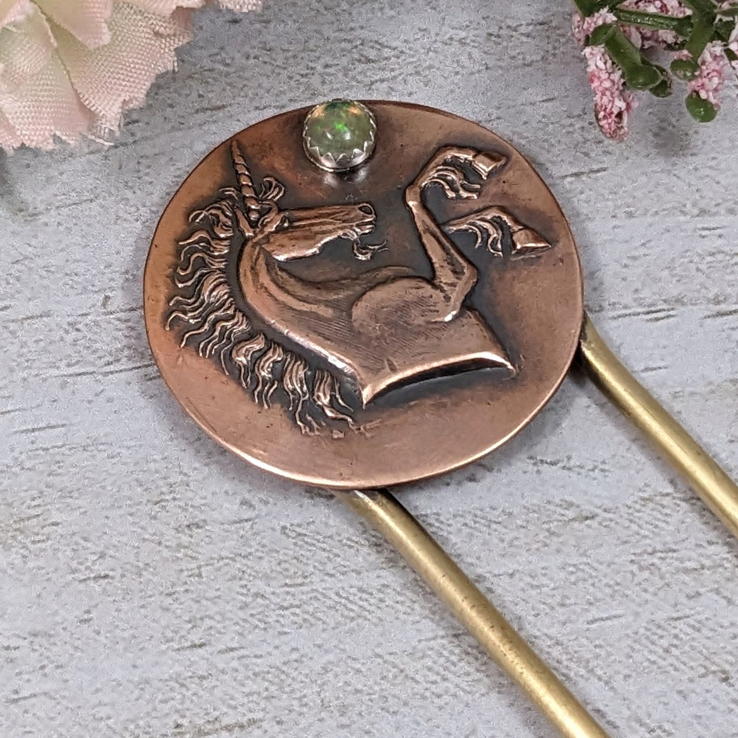 Brass hair fork with a copper design at the top. The copper is round with a raised impression of a unicorn. You can see the beard, detailed hair in the mane and on the ankles. The horn is spiraled. Above the nose there is a round gemstone meant to represent the moon.