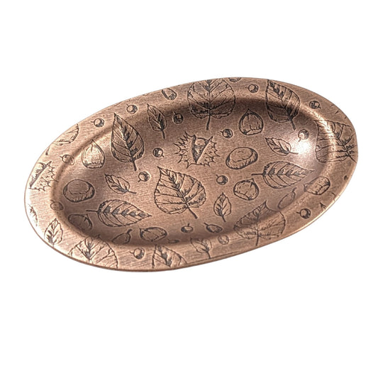 Oval copper ring dish with a raised edge. Pattern on dish is leaves, acorns, and other fall tree nuts.