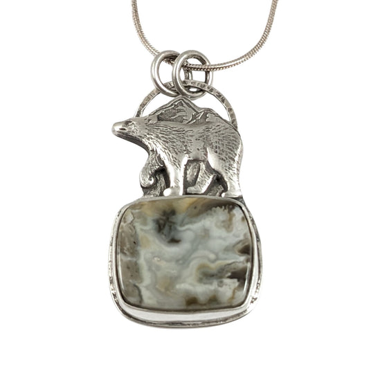 Sterling silver pendant. The top half is a bear walking against a background of mountains, the bottom half is a cabochon that looks like river rapids.