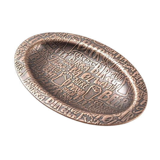 An oval copper ring dish with a raised edge. The dish has an impressed pattern made up of the word beer in many languages.