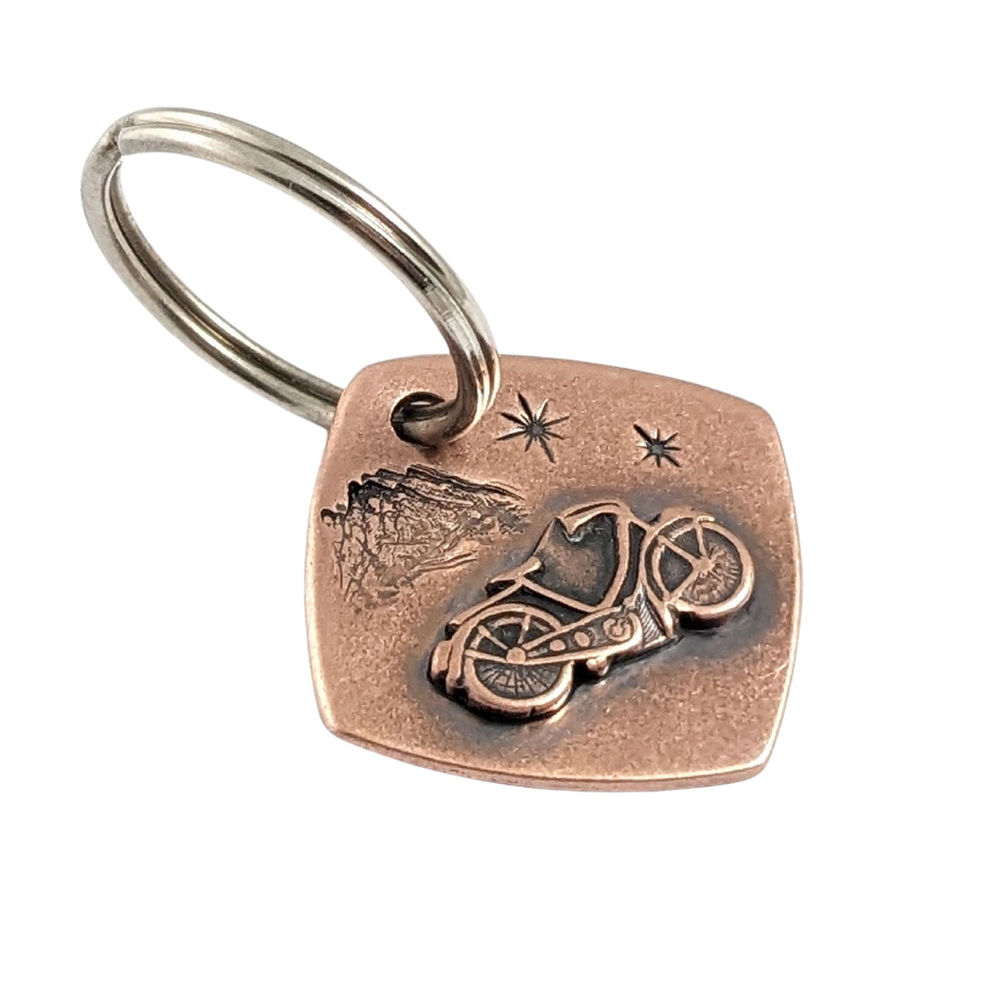 copper keychain with raised detailed bicycle. background is mountains and twinkling stars