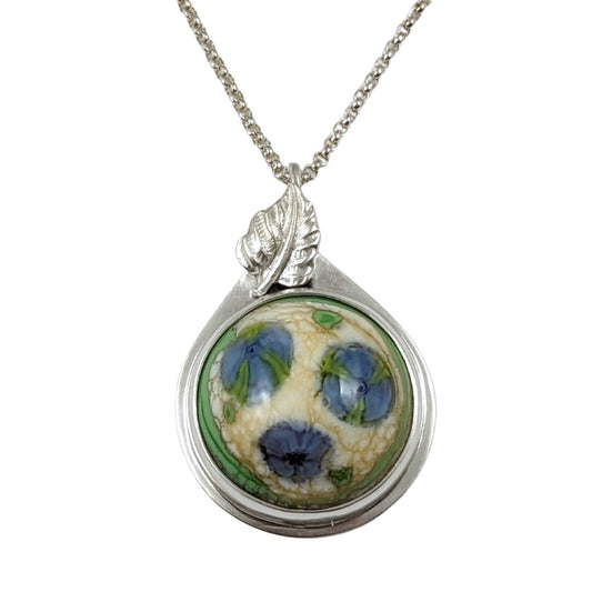 Sterling silver pendant. The stone is a round lampwork glass cabochon with three blue flowers. The flowers have bits of green in them. The background is pale yellow and there is a border of green. Above the stone and partially hiding the bale is a silver leaf.