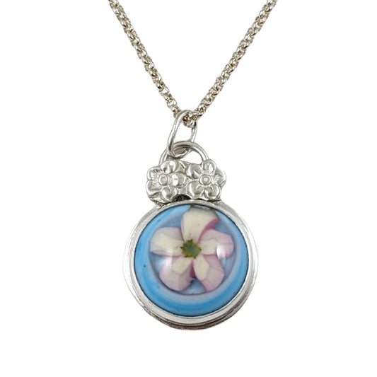 Sterling silver pendant with a glass cabochon. The cabochon has a white five petal flower and a blue background. Above the cabochon setting there are two small five petal flowers made from sterling silver, then a small bail.