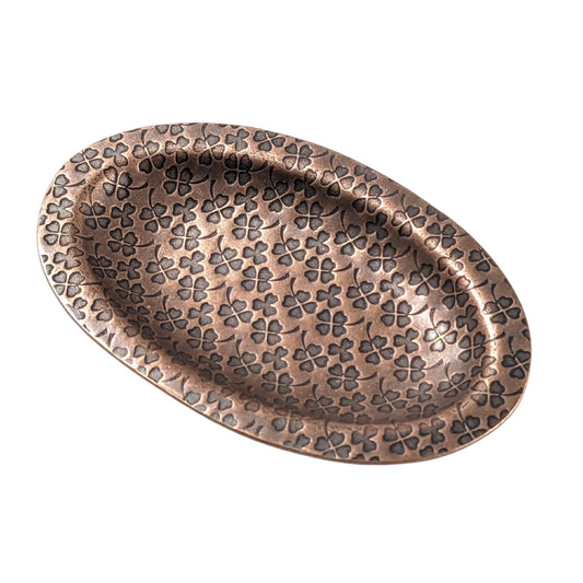 An oval copper ring dish with a raised edge. The dish has an impressed pattern of four leaf clovers.