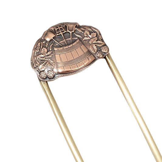 brass hair fork with a dimensional design in copper. The design has a wooden barrel in the middle, filled with three long handled garden tools. Surrounding the barrel are leaves and strawberries.
