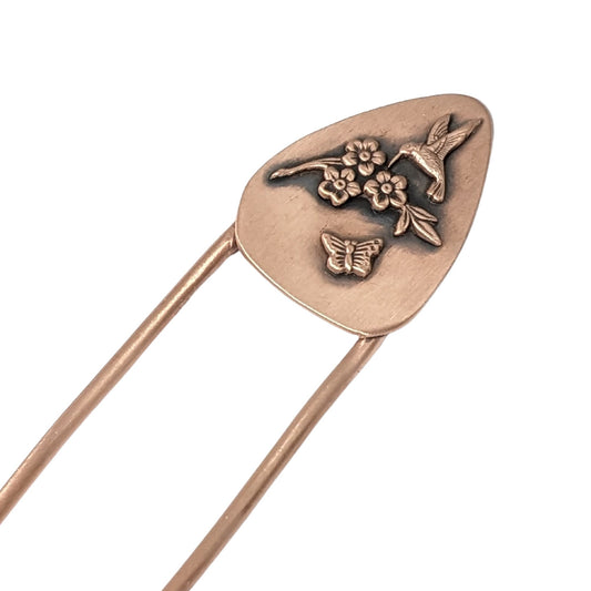 Copper hair fork with a design added to the top. The design is copper and is roughly egg-shaped. On the design are three-dimensional flowers, a hummingbird, and a butterfly.