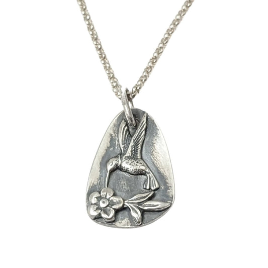 Sterling silver pendant in a freeform oval shape. On the pendant there is a silver hummingbird in flight, hovering over a single flower with three leaves.