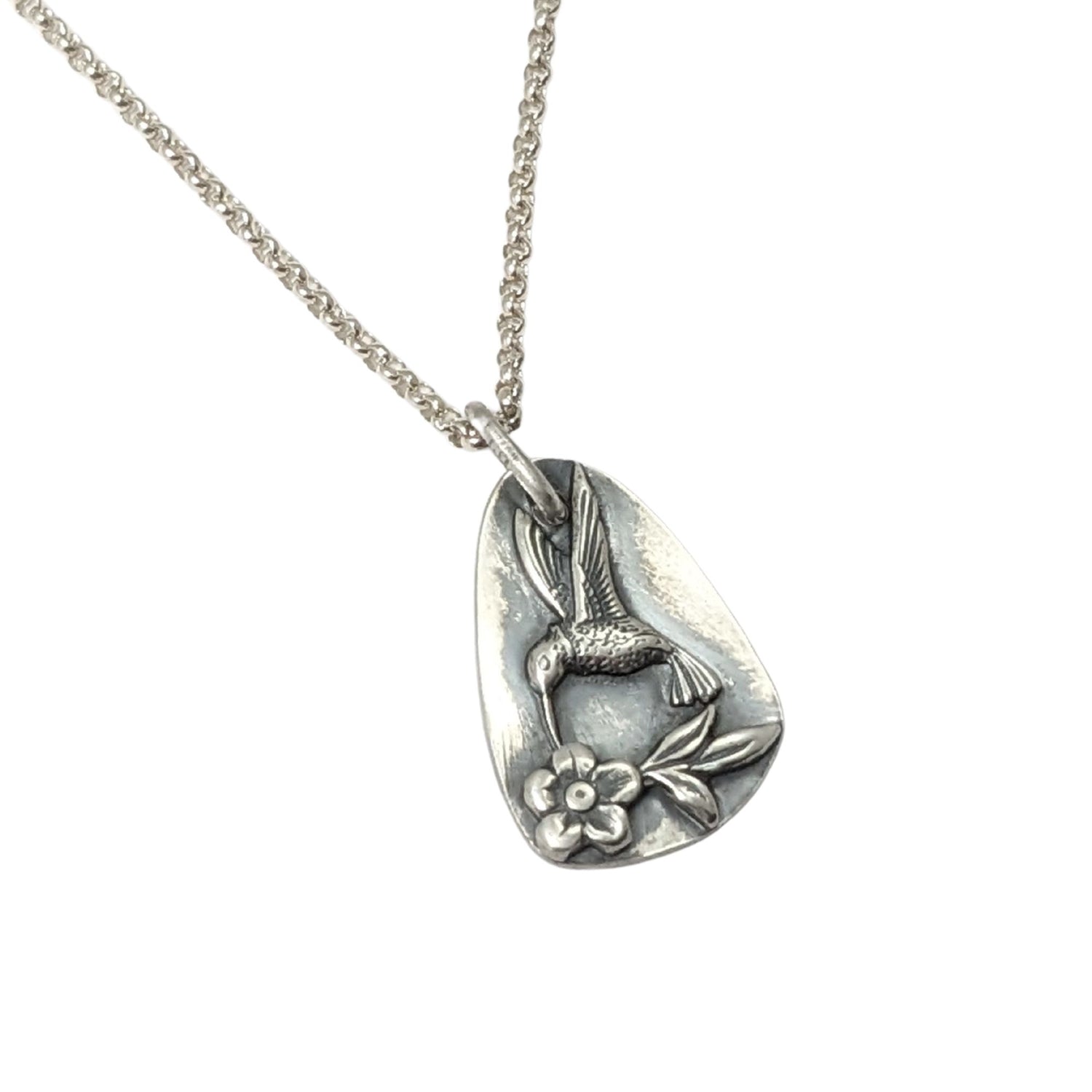 Sterling silver pendant in a freeform oval shape. On the pendant there is a silver hummingbird in flight, hovering over a single flower with three leaves.
