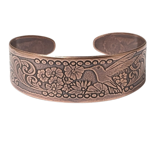 Tapered copper cuff bracelet. Pattern across middle is a hummingbird surrounded by flowers. At each end of the cuff there is a smaller hummingbird and flower.