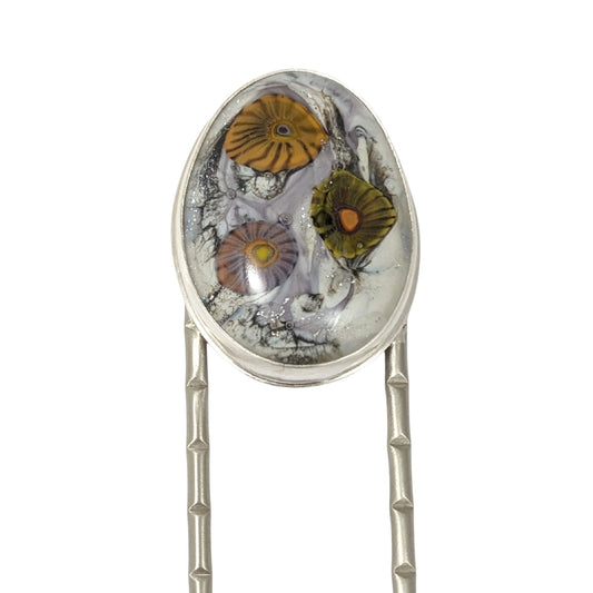 Lampwork glass oval with three abstract flowers in yellow, green, and pale purple set on a nickel hair fork.