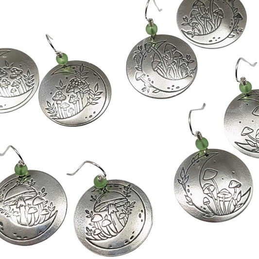 A set of domed sterling silver round drop earrings featuring an engraved mushroom and crescent moon design, accented with small green beads and silver ear wires.