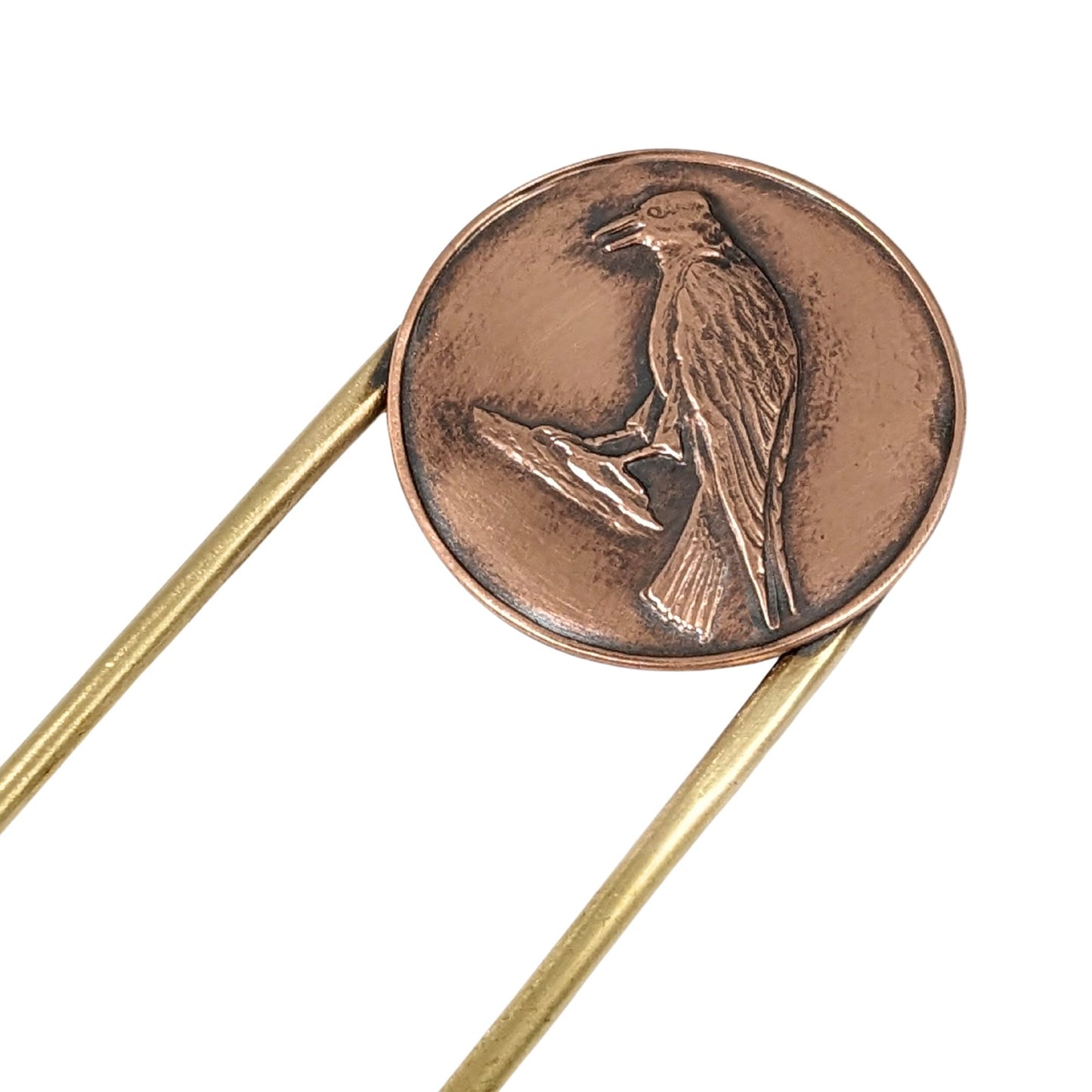 Brass hair fork with a design element on the end. The design is round copper with a raised image of a raven standing on a rock.