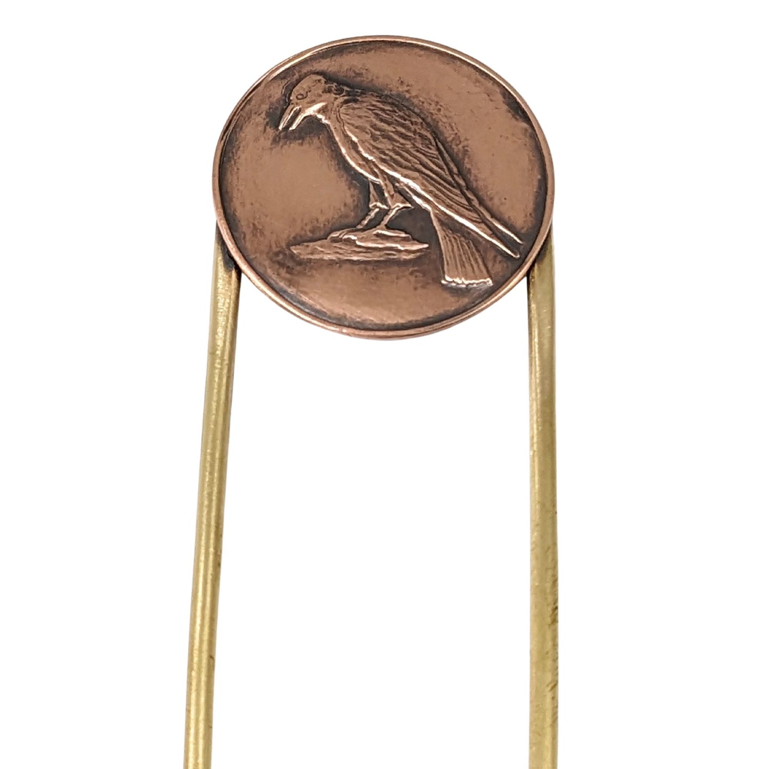 Brass hair fork with a design element on the end. The design is round copper with a raised image of a raven standing on a rock.