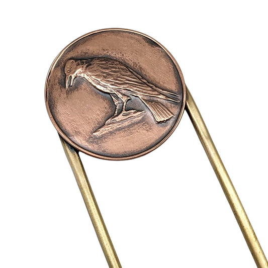 Brass hair fork with a design element on the end. The design is round copper with a raised image of a raven standing on a rock.