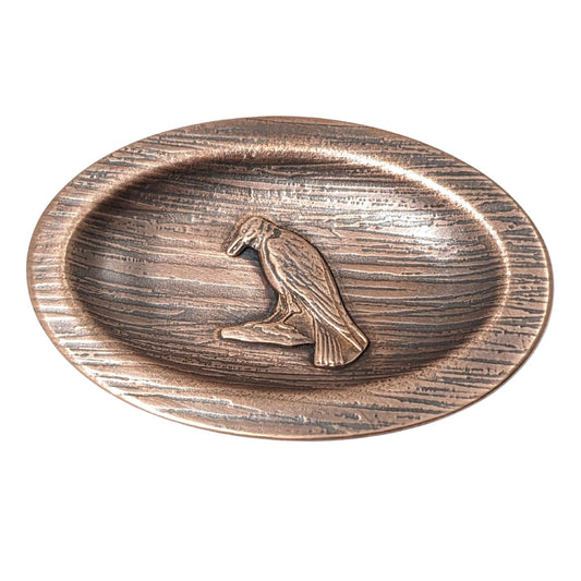 Oval copper ring dish with a raised lip edge. The copper is textured to resemble wood, in the middle is a raised image of a raven perched on a rock.