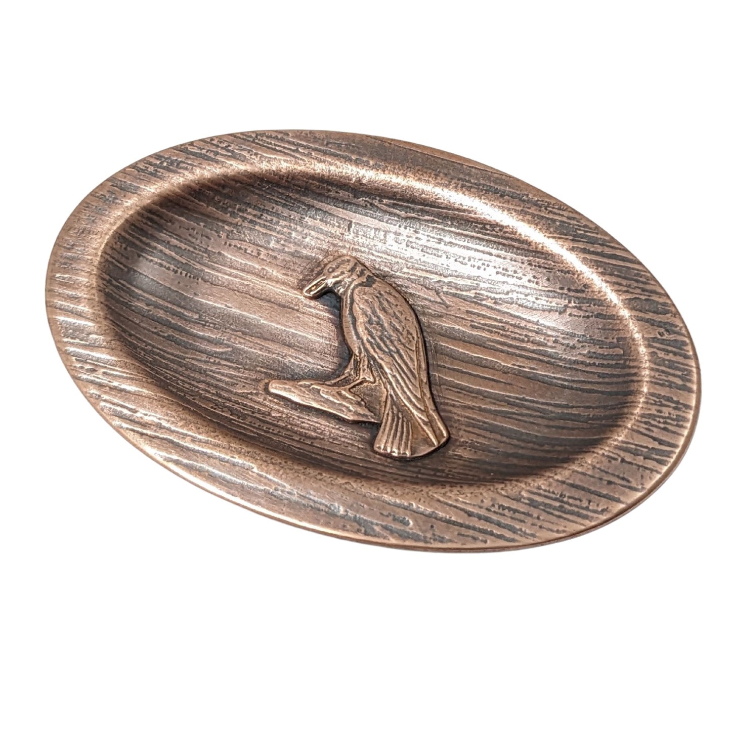 Oval copper ring dish with a raised lip edge. The copper is textured to resemble wood, in the middle is a raised image of a raven perched on a rock.