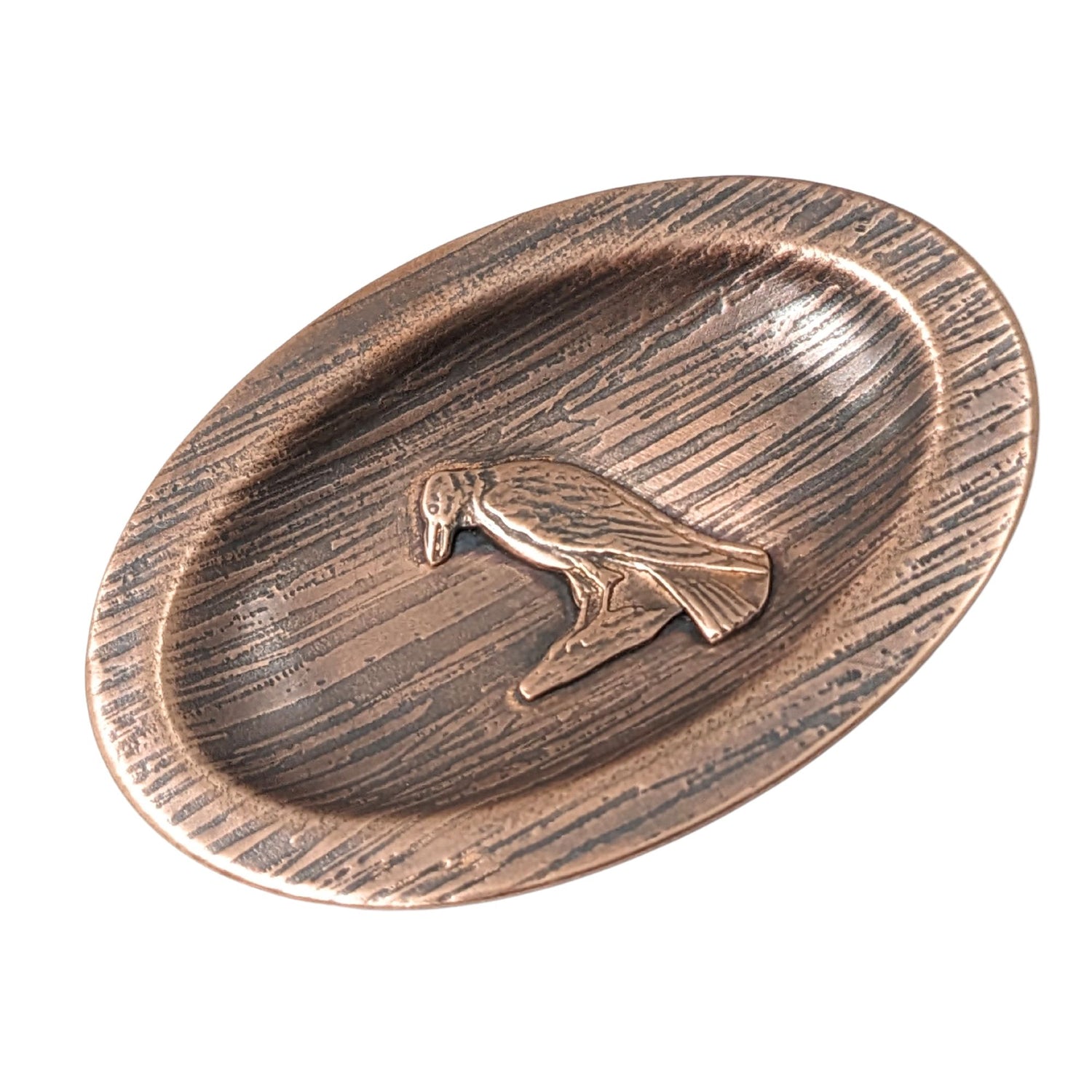 Oval copper ring dish with a raised lip edge. The copper is textured to resemble wood, in the middle is a raised image of a raven perched on a rock.