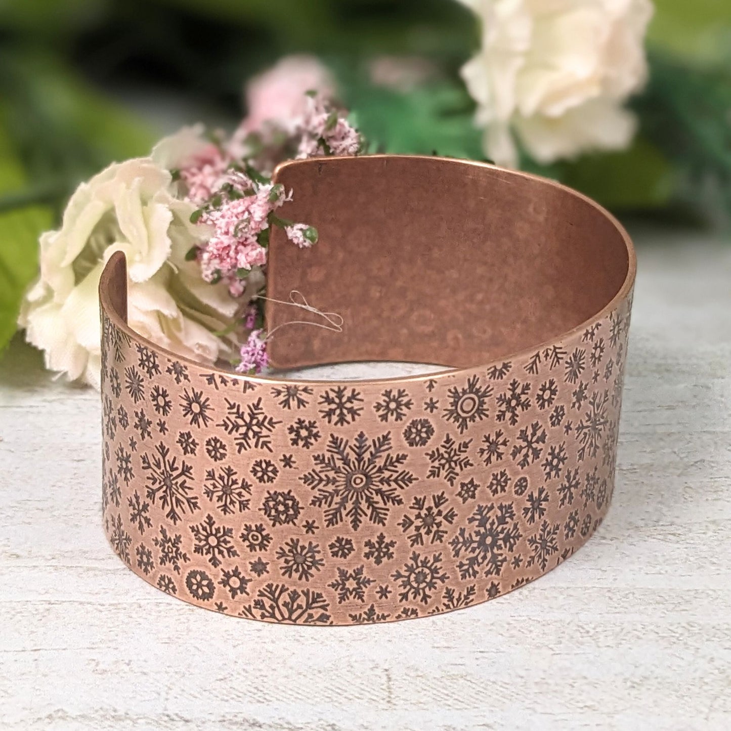 inch wide copper cuff bracelet with impressed design of different snowflake shapes. the impressions are oxidized, or blackened, to enhance the design. staged with flowers and leaves in the background