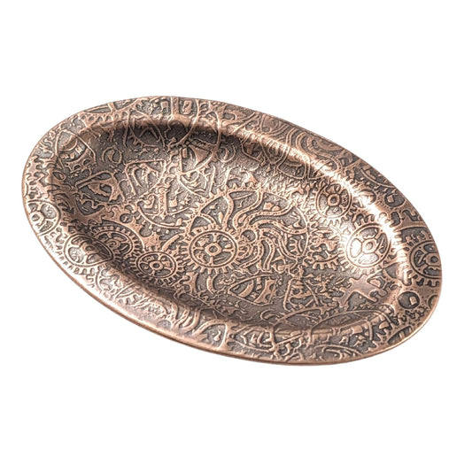 An oval copper ring dish with a raised edge. The dish has an impressed pattern of steampunk style gears and cogs.
