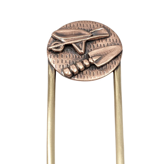 Copper hair fork with a round design on the end of the fork. The design is a round piece of copper textured to resemble seeds. Soldered to the round is a copper wheelbarrow and a copper hand trowel. The fork is brass.