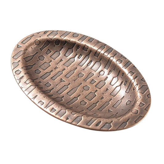 An oval copper ring dish with a raised edge. The dish has an impressed designs of wine and cocktail glasses plus wine and liquor bottles.