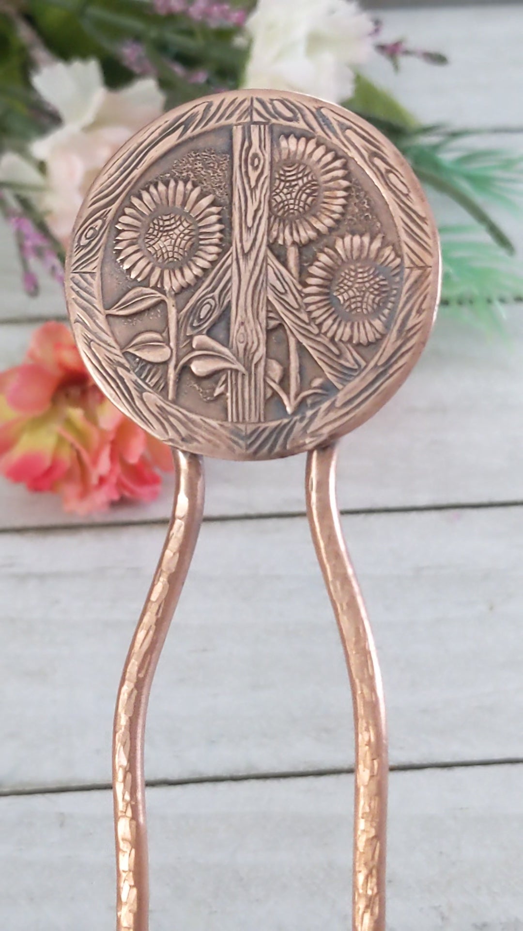 Video of Copper hair fork with a large round design at top of sunflowers and a peace sign. The peace sign has a wood texture. Three sunflowers grow through the wood sign. There's a lot of detail - veins on the leaves, you can see the sunflower seeds and petals. On a copper fork with a hammered finish.