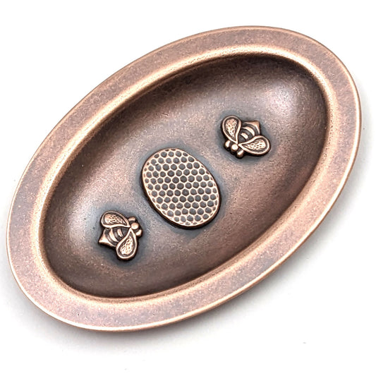 Oval copper ring dish. In the middle is a three dimensional oval honeycomb pattern. On either side of the honeycomb is a three dimensional bee. The dish has a small lip around the edge.