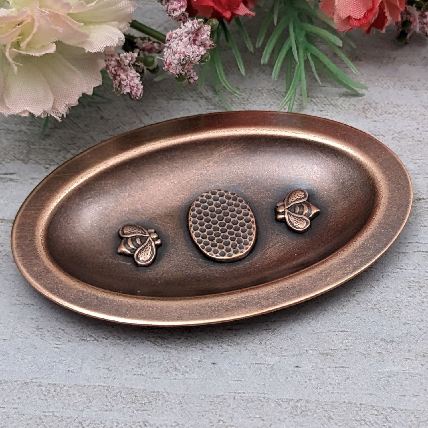 Oval copper ring dish. In the middle is a three dimensional oval honeycomb pattern. On either side of the honeycomb is a three dimensional bee. The dish has a small lip around the edge.