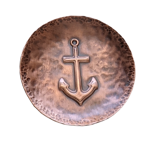 Copper ring or jewelry or trinket dish with a raised impression of a boat anchor. The edged of the dish are curved up slightly, the copper metal has a hammered texture, and the dish is oxidized to enhance the detail.