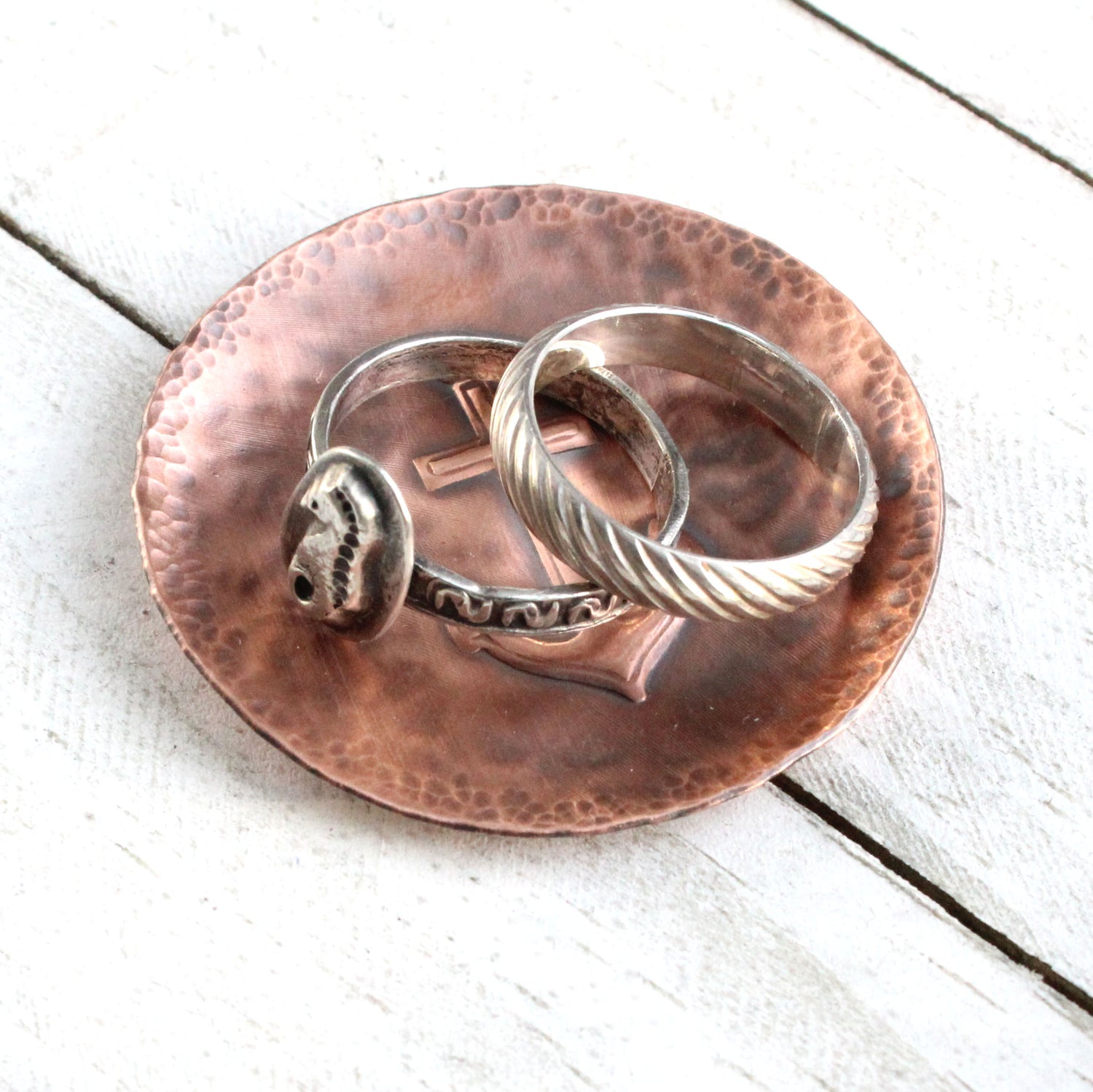 Copper ring or jewelry or trinket dish with a raised impression of a boat anchor. The edged of the dish are curved up slightly, the copper metal has a hammered texture, and the dish is oxidized to enhance the detail. Shown with rings set on the dish.