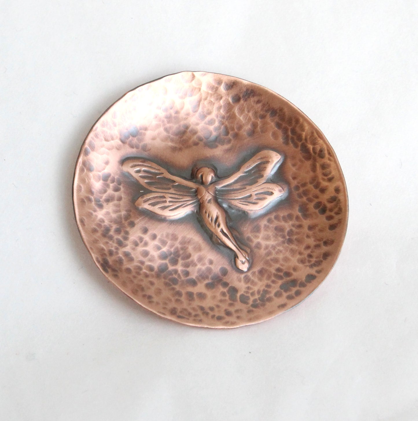 Small copper copper dish for rings, jewelry, trinkets. In middle there is a raised image of a fairy in flight. The background is dappled hammer texture, the sides are raised to form a shallow bowl.