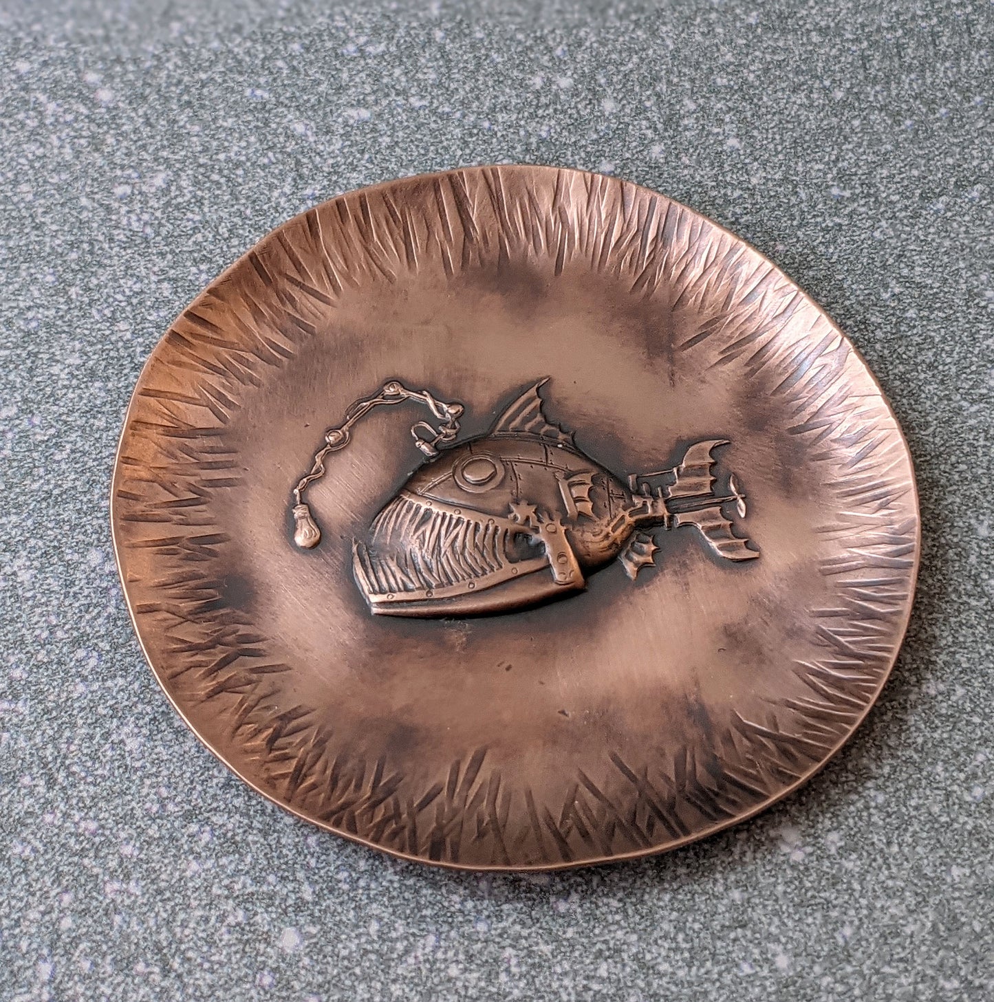 A copper dish with a raised impression of an anglerfish. This is a classic steampunk design inspired by the works of Jules Verne. The dish is used to hold rings and jewelry. The edges are slightly raised to create a bowl shape and have hammered lines design around the edges. The piece is oxidized to enhance the details.