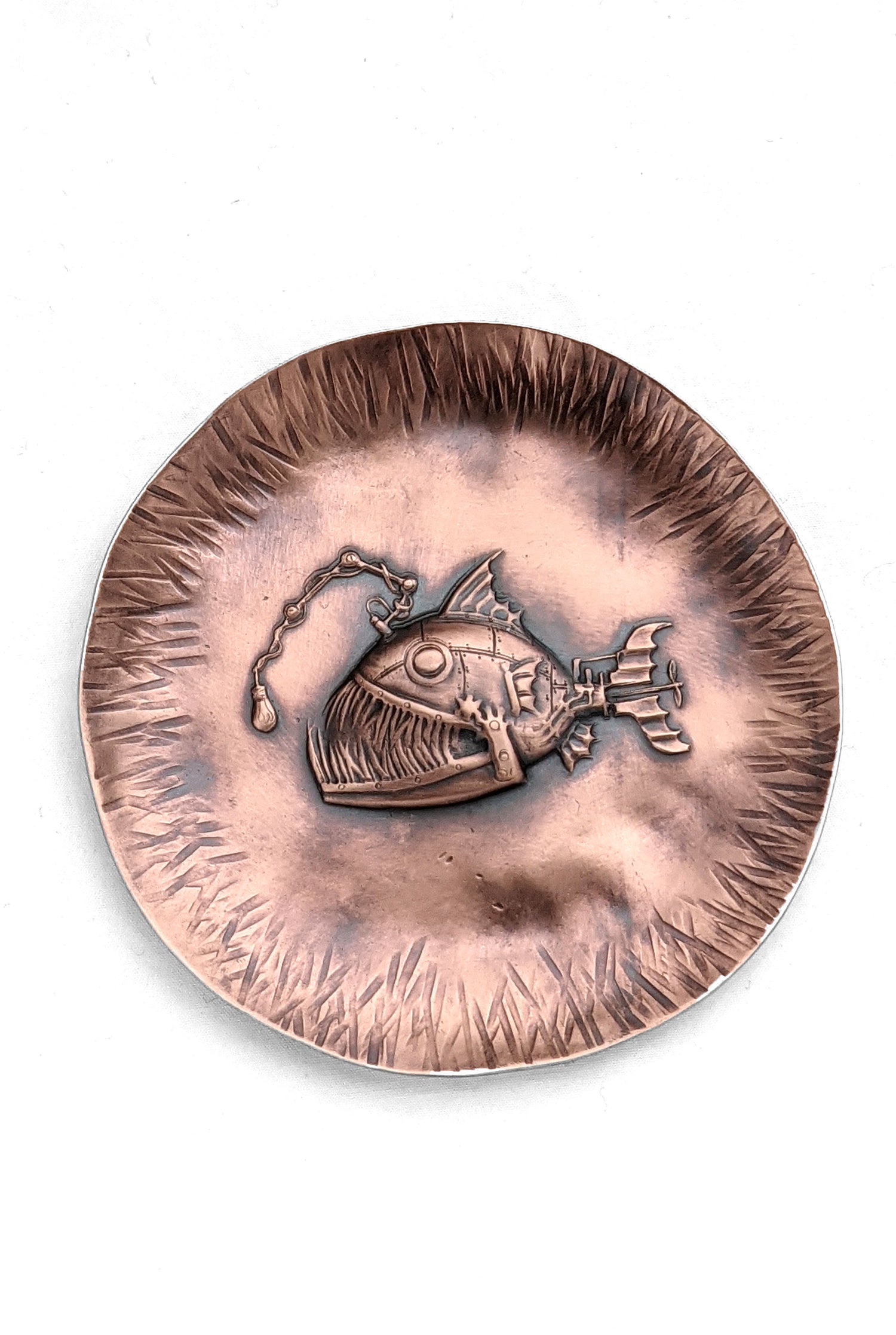 A copper dish with a raised impression of an anglerfish. This is a classic steampunk design inspired by the works of Jules Verne. The dish is used to hold rings and jewelry. The edges are slightly raised to create a bowl shape and have hammered lines design around the edges. The piece is oxidized to enhance the details.