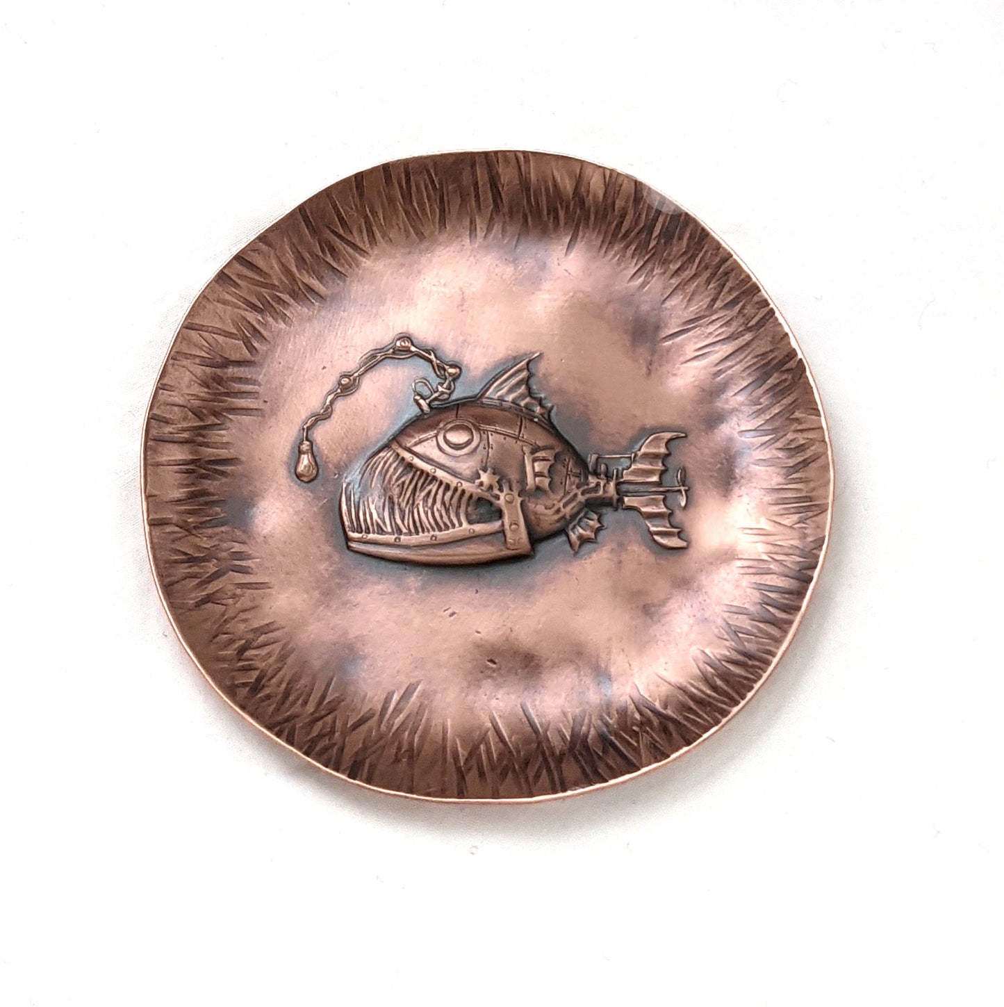 A copper dish with a raised impression of an anglerfish. This is a classic steampunk design inspired by the works of Jules Verne. The dish is used to hold rings and jewelry. The edges are slightly raised to create a bowl shape and have hammered lines design around the edges. The piece is oxidized to enhance the details.