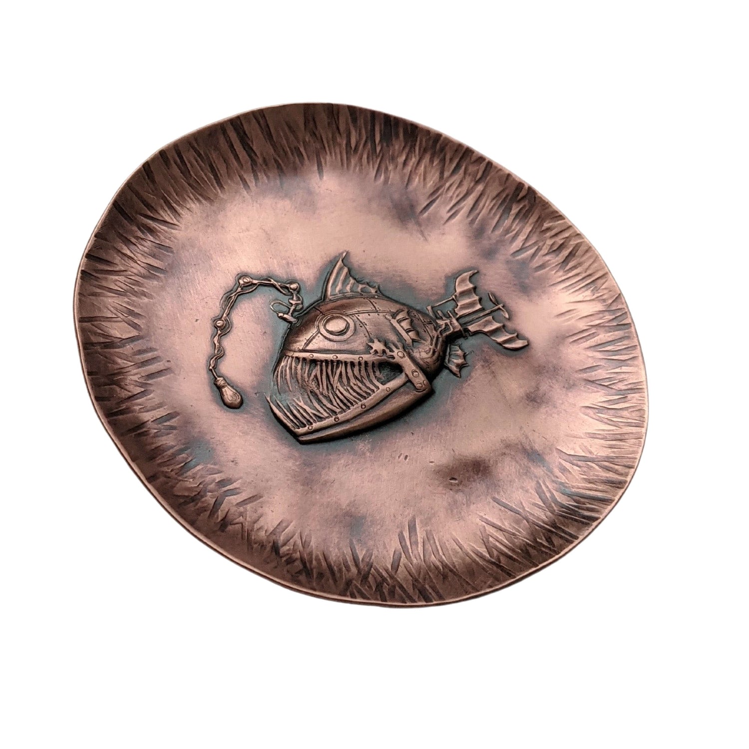 A copper dish with a raised impression of an anglerfish. This is a classic steampunk design inspired by the works of Jules Verne. The dish is used to hold rings and jewelry. The edges are slightly raised to create a bowl shape and have hammered lines design around the edges. The piece is oxidized to enhance the details.