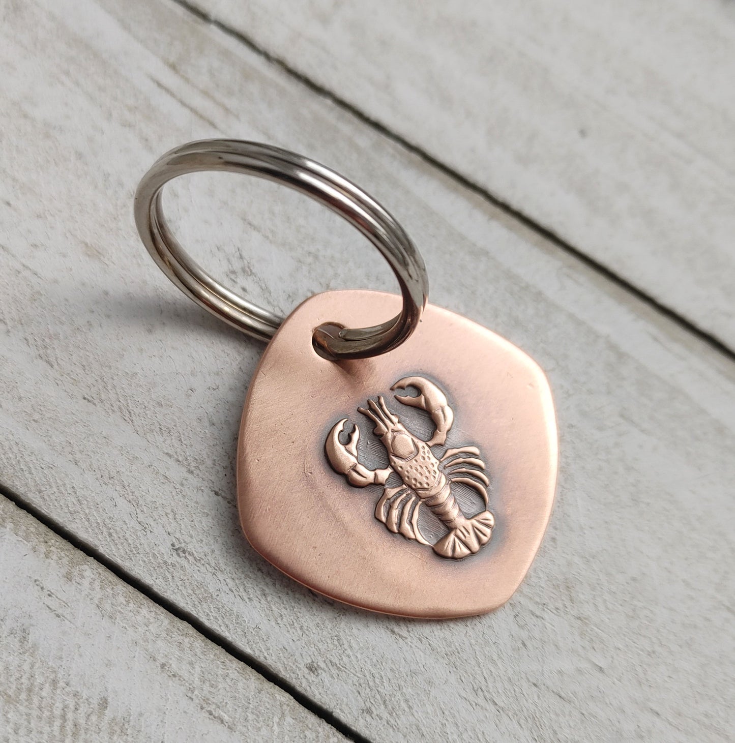 Copper keychain with a detailed raised impression of a lobster. The claws, legs, and tail are all clearly defined. The keychain is a rounded square shape.