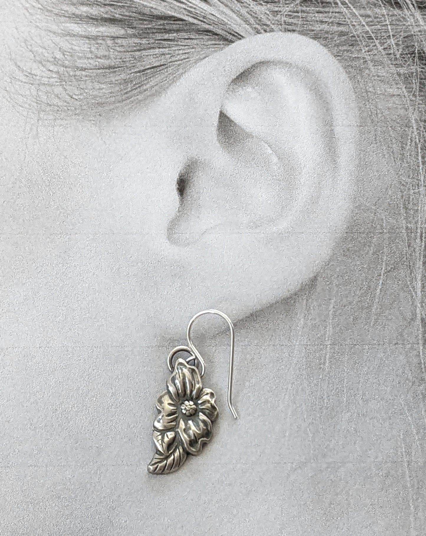Stering silver dogwood flower earrings. The Design is asymmetric, the left earring has a leaf above the flower and the right earring has a leaf below the flower. Shown modeled on picture of a womans ear