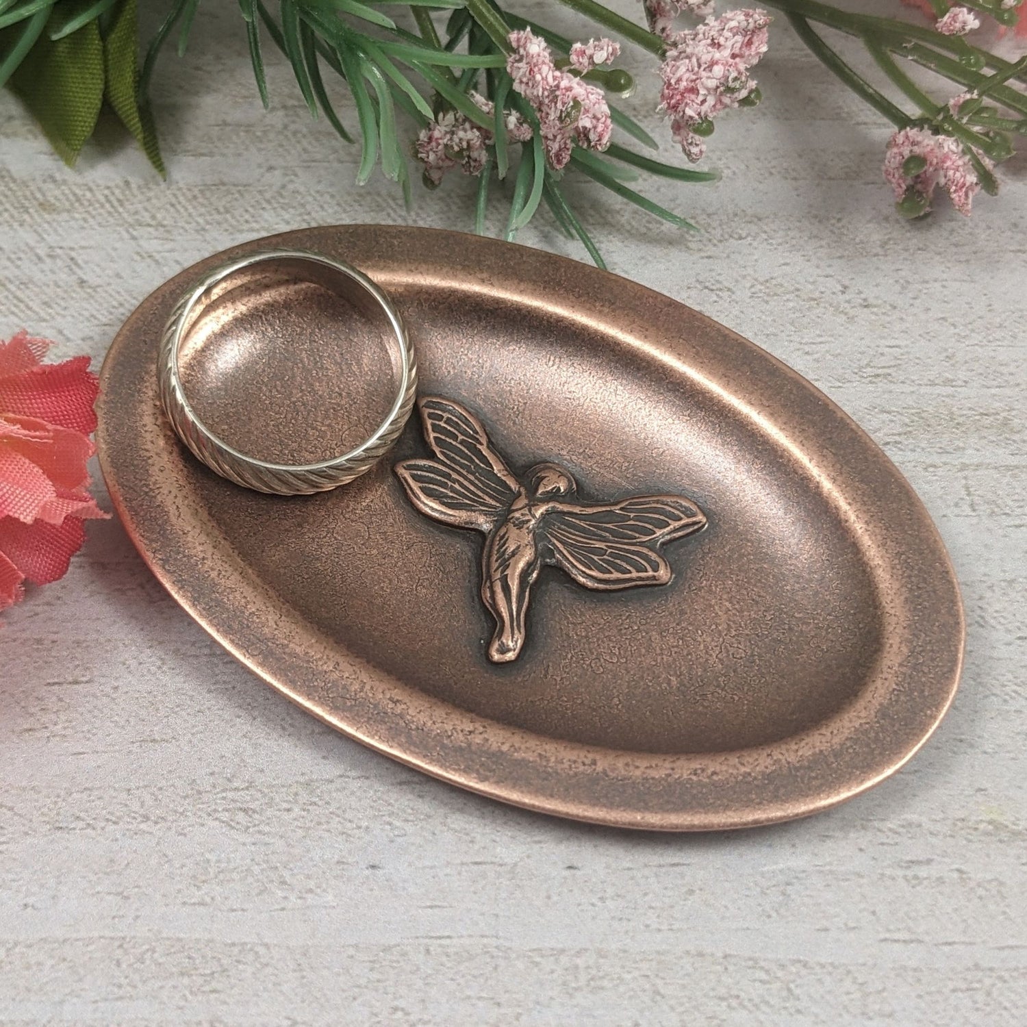 Oval copper ring dish. In the middle is a three dimensional fairy in flight. Her wings have detailed webbing pattern and you can see the draping on her dress.  The dish has a small lip around the edge. Staged with a ring in the dish and flowers in the background.
