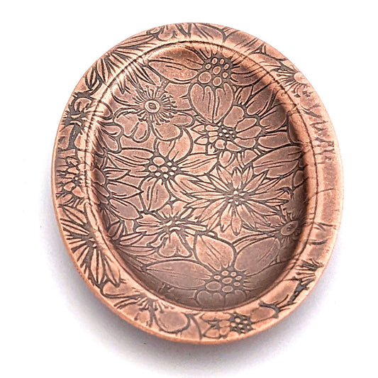 Copper oval ring dish with repeating pattern of assortment of flowers.  Dish is 2 inches by three inches with a raised lip.
