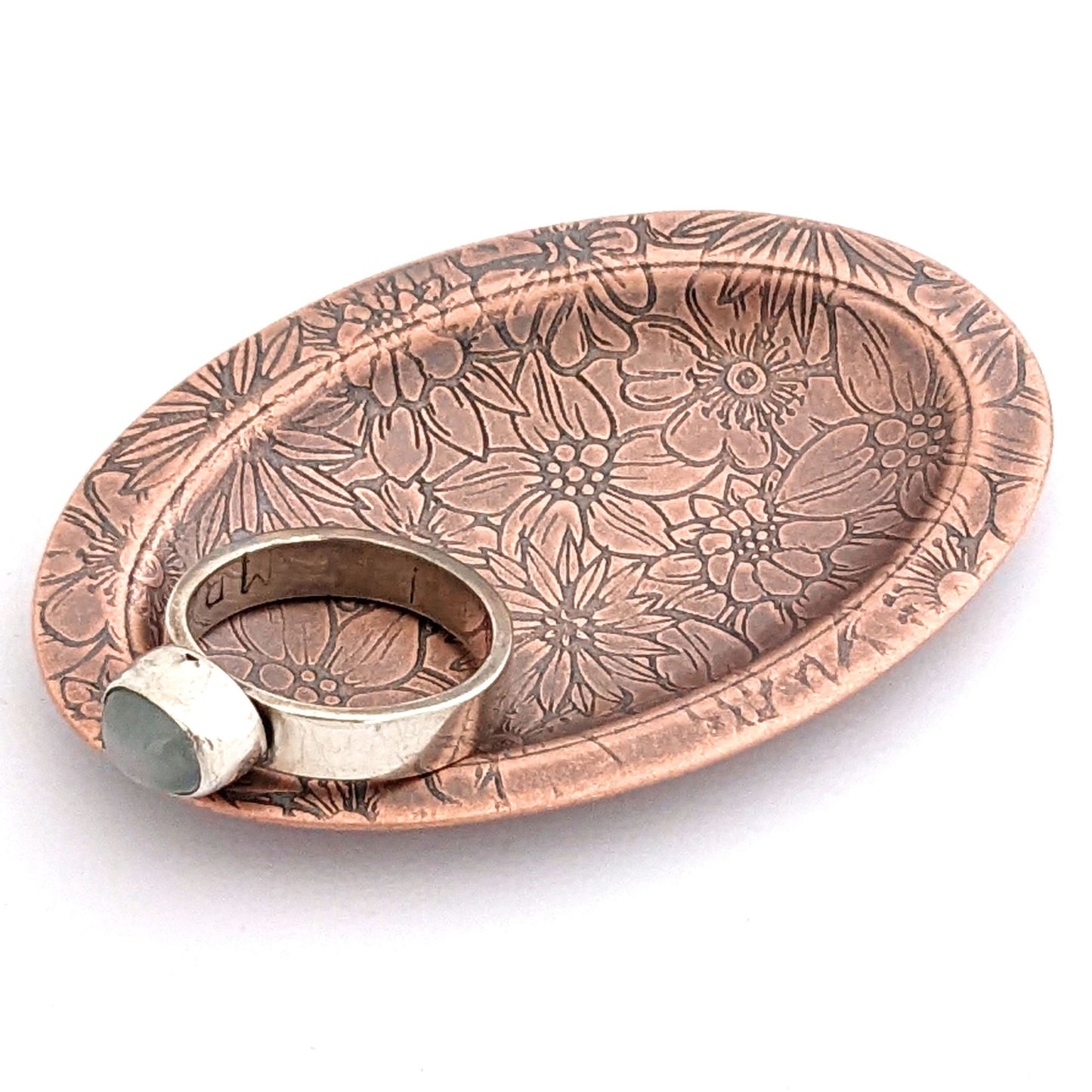 Copper oval ring dish with repeating pattern of assortment of flowers.  Dish is 2 inches by three inches with a raised lip.
